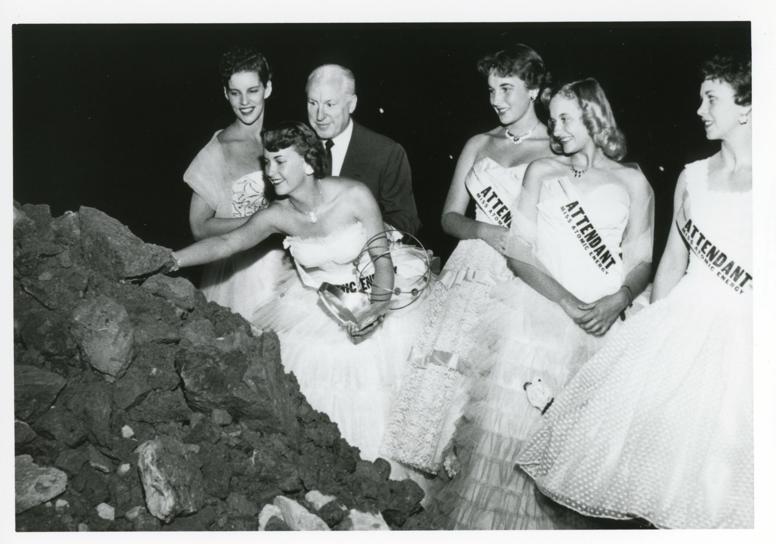 Beauty queens picking up a piece of uranium ore out of a pile.